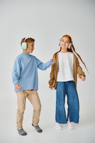 joyous little boy and girl in sweatshirt and cardigan with headphones on gray backdrop, fashion