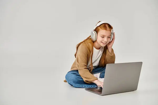 stock image pretty cheerful little girl in winter trendy attire on floor with headset looking at laptop, fashion