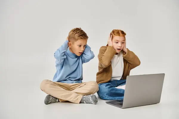 stock image two scared trendy little children looking at laptop and closing ears with hands, fashion concept
