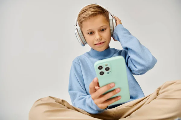 stock image little boy in trendy sweatshirt on floor with headset looking at his phone, smiling slightly