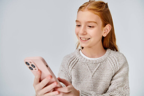 cheerful preteen girl with red hair looking joyfully at her phone on gray backdrop, fashion concept