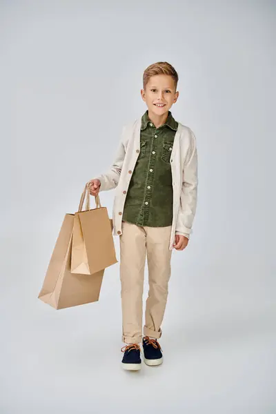 Stock image joyous little boy in stylish casual cardigan posing on gray backdrop with present bags, fashion