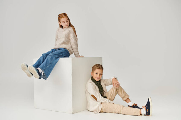 red haired little girl sitting on cube and looking at smiley preteen boy sitting on floor, fashion