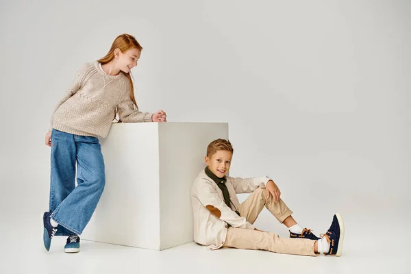 jolly preteen girl standing next to white cube and looking at little boy sitting on floor, fashion