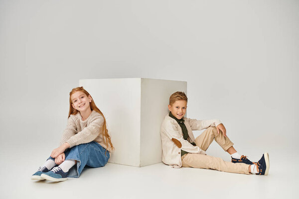 happy preadolescent children sitting on floor next to white cube and smiling at camera, fashion