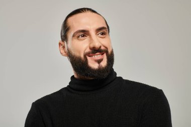 portrait of happy and handsome arabic man with beard posing in black turtleneck on grey backdrop clipart