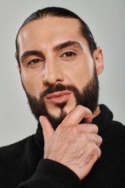 portrait of good looking arabic man in turtleneck touching beard and posing on grey backdrop clipart