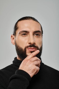 portrait of good looking arabic man in turtleneck touching beard while thinking on grey backdrop clipart