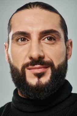 Siyah balıkçı yaka kazaklı, gri arka planda poz veren gülümseyen sakallı Arap adamın yakın plan çekimi.