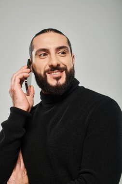 Sakallı neşeli Arap adam gülümsüyor ve gri arka planda akıllı telefondan telefon alıyor.
