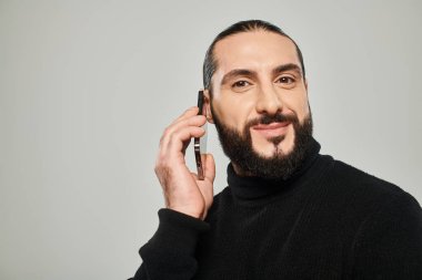 happy arabic man with beard smiling and having phone call on smartphone on grey background clipart