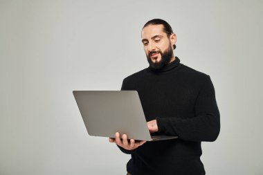 Siyah balıkçı yaka kazaklı yakışıklı ve sakallı bir adam gri arka planda dizüstü bilgisayarla dikiliyor.