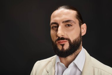 Beyaz gömlekli ve ceketli yakışıklı ve sakallı Arap adam siyah arka planda kameraya bakıyor.