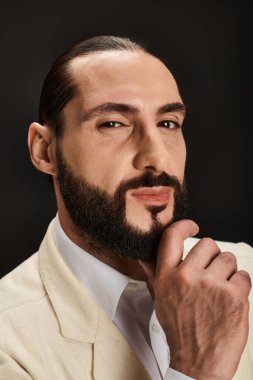 Beyaz gömlekli, spor ceketli, sakallı, siyah arka planda kameraya bakan Arap adamın portresi.