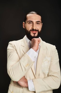 Beyaz gömlekli ve ceketli neşeli sakallı Arap adam siyah arka planda kameraya bakıyor.
