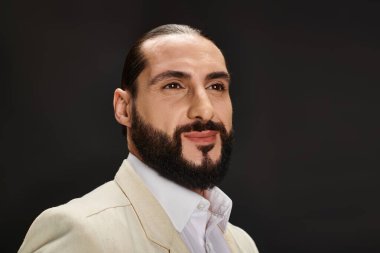 Beyaz gömlekli ve siyah arkaplanda poz veren zarif ceketli neşeli sakallı Arap adamın portresi.