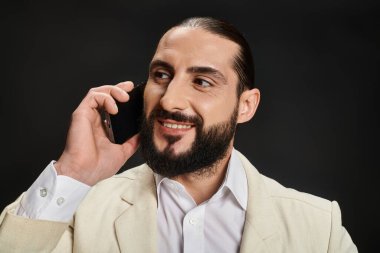 Beyaz gömlekli ve ceketli neşeli ve sakallı Arap adam siyah arka planda akıllı telefondan konuşuyor.