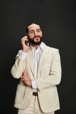 Beyaz gömlekli ve ceketli neşeli ve sakallı Arap adam siyah arka planda akıllı telefondan konuşuyor.