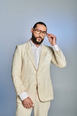 Gözlüklü ve sakallı bir adam gri arka planda beyaz giyinmiş, resmi giyinmiş.