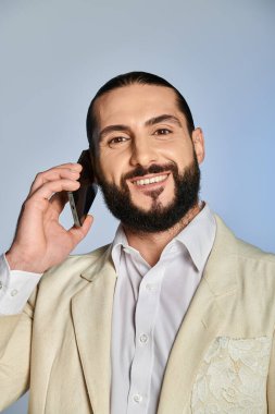 Gri arka planda akıllı telefondan konuşan şık giyimli mutlu sakallı Arap adam.