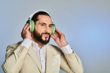 Zarif görünümlü sakallı Arap adam gri arka planda kablosuz kulaklıkla müzik dinliyor.