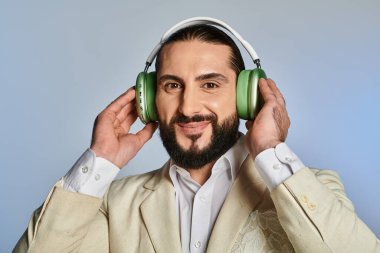 Şık giyinmiş mutlu sakallı adam gri arka planda kablosuz kulaklıkla müzik dinliyor.