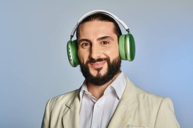 Şık giyinmiş mutlu sakallı adam gri arka planda kablosuz kulaklıkla müzik dinliyor.