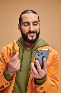 Mutlu adam baklava tutuyor ve bej arka planda akıllı telefon kullanıyor. Geleneksel Orta Doğu tatlıları.