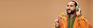 cheerful man in headphones holding honey baklava on beige background, middle eastern dessert banner clipart