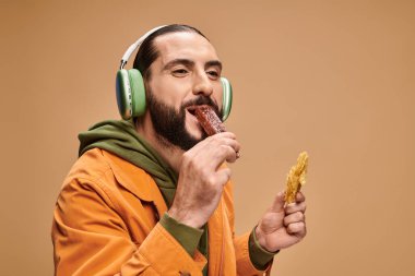 happy arabic man in headphones holding honey baklava and biting churchkhela on beige backdrop clipart