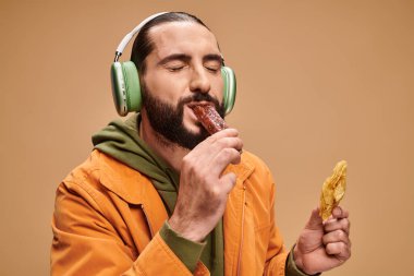 happy arabic man in headphones holding honey baklava and biting churchkhela on beige backdrop clipart