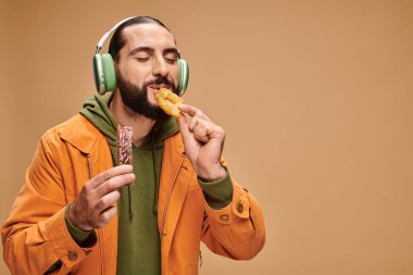 Kulaklıklı neşeli bir adam bej arka planda ballı baklava yiyor.