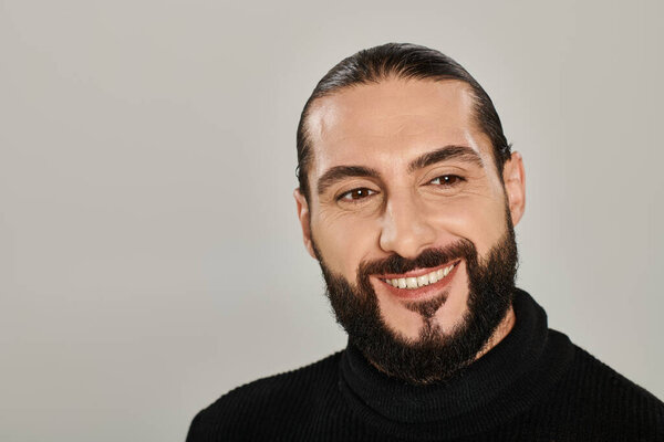 portrait of happy good looking arabic man with beard posing in black turtleneck on grey backdrop