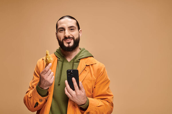happy man holding baklava and using smartphone on beige backdrop, traditional middle eastern dessert