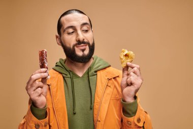 joyful bearded man smiling and holding baklava and cevizli sucuk on beige backdrop, turkish delights clipart