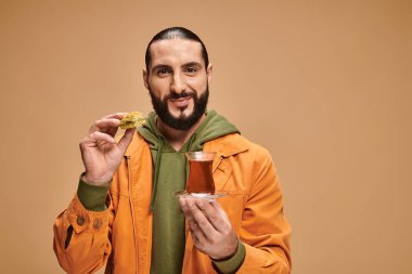 Geleneksel cam bardakta Türk çayı ve bej renkli baklava tutan günlük kıyafetli mutlu sakallı bir adam.