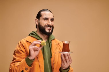 Geleneksel cam bardakta Türk çayı ve bej renkli baklava tutan günlük kıyafetli mutlu sakallı bir adam.