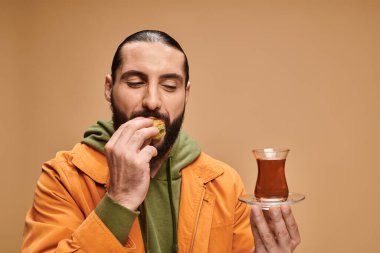 Mutlu sakallı adam geleneksel cam fincanda Türk çayı tutuyor ve bej rengi üzerinde lezzetli baklava yiyor.