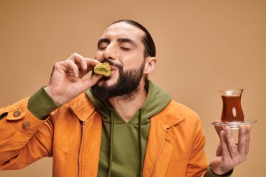 Mutlu sakallı adam geleneksel cam fincanda Türk çayı tutuyor ve bej rengi üzerinde lezzetli baklava yiyor.