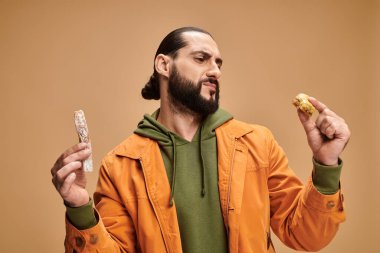 Kafası karışmış Arap sakallı adam elinde baklava ve cevizli sucuk tutuyor bej arka planda, Türk lezzetleri