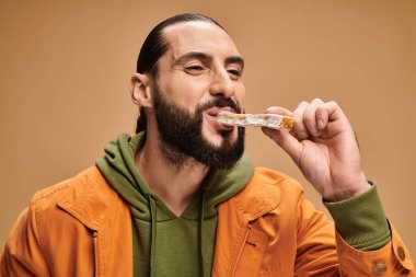 Neşeli ve sakallı Arap adam bej arka planda lezzetli cevizli sucuk yiyor.