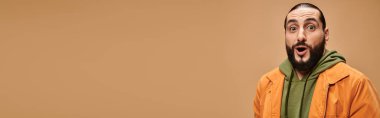 Günlük giysiler içinde sakallı, bej arka planda kameraya bakan Arap bir adam şaşırdı.