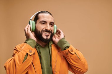 Sakallı neşeli orta doğulu adam bej arka planda kablosuz kulaklıkla müzik dinliyor.