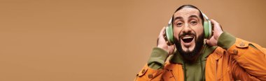 Sakallı neşeli orta doğulu adam bej, pankartta kablosuz kulaklıkla müzik dinliyor.