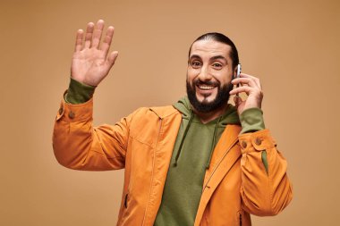 Dost canlısı, sakallı, bej arka planda akıllı telefondan konuşan, el sallayan orta doğulu bir adam.