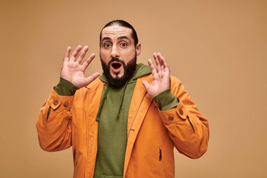 Şaşırmış, sakallı, ağzı açık, bej arka planda el kol hareketi yapan orta doğulu bir adam.