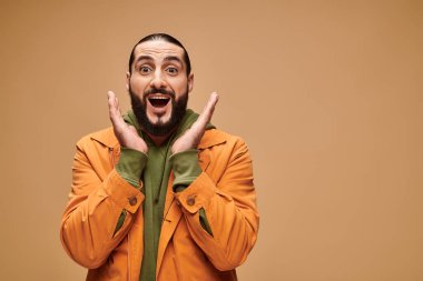 Şaşırmış Orta Doğulu sakallı ve ağzı açık bir adam bej arka planda el kol hareketi yapıyor.