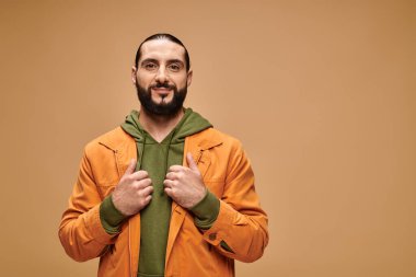 Memnun orta doğulu sakallı bir adam bej arka planda günlük giysiler içinde kameraya bakıyor.