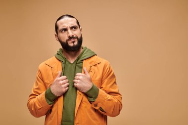 Sakallı, bej arka planda sıradan giyinmiş dalgın bir orta doğulu adam başka tarafa bakıyor.