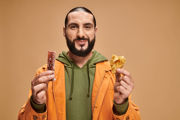 happy bearded man smiling and holding baklava and cevizli sucuk on beige backdrop, turkish delights